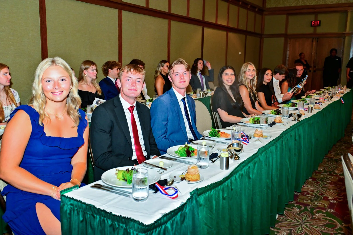 A group of young adults dressed in formal attire seated at a banquet table during a formal event or celebration.