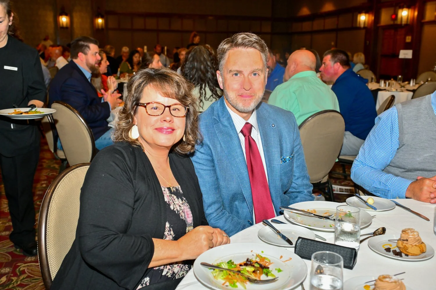 Two people, a woman with glasses and a man in a blue suit, sitting at a dining table during a formal event.