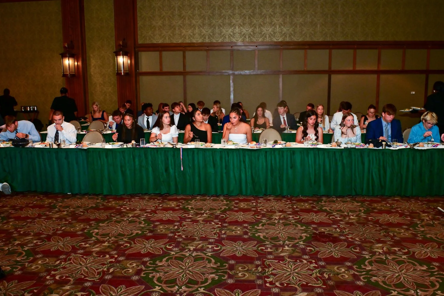 A group of young people dressed formally, sitting at a long banquet table in a large, decorated room.