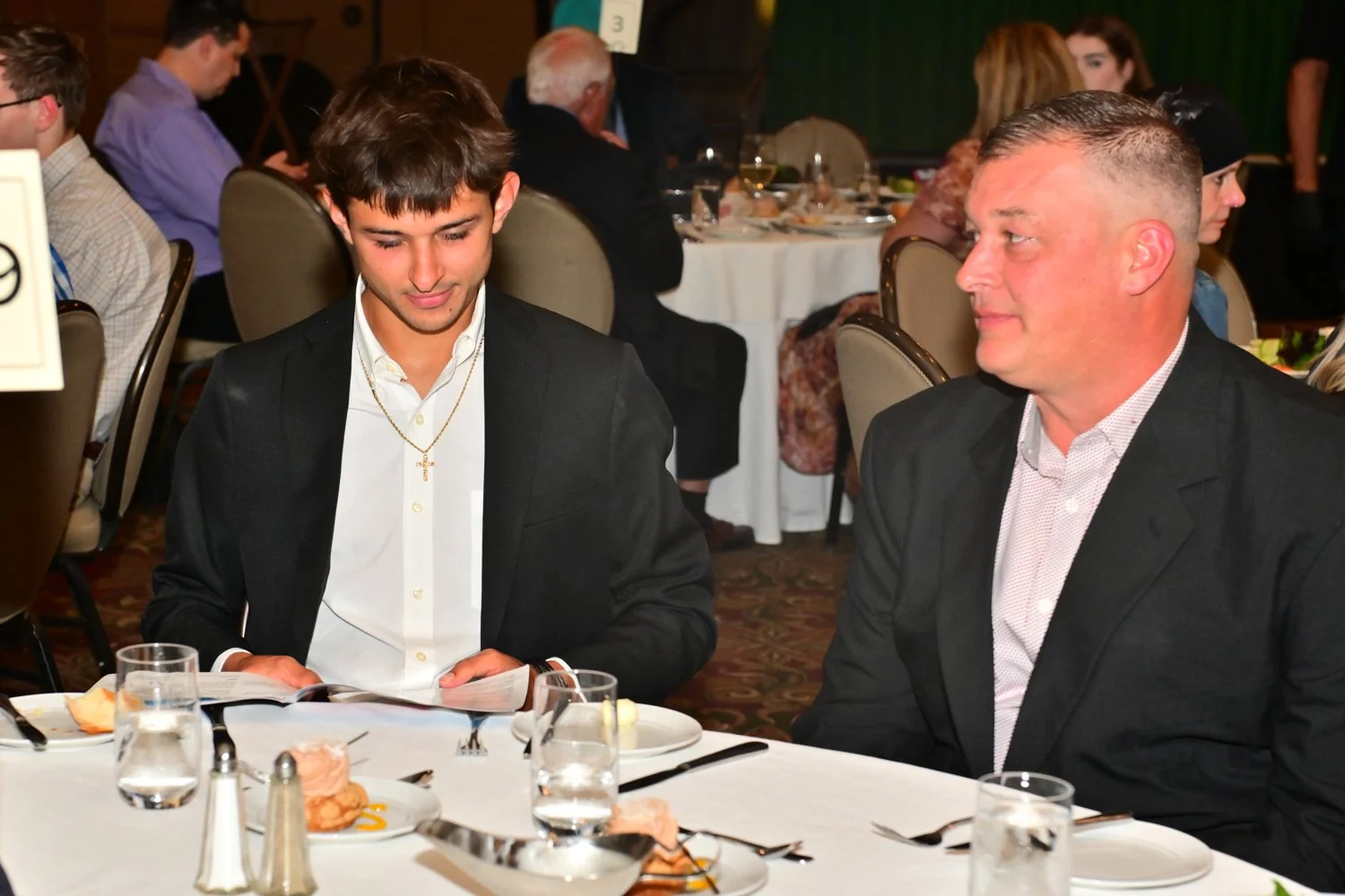 Two men dressed in suits sitting at a dining table in a banquet hall. The younger man on the left is looking at a menu, wearing a black blazer, white shirt, and a gold cross necklace. The older man on the right is looking at the younger man, wearing 