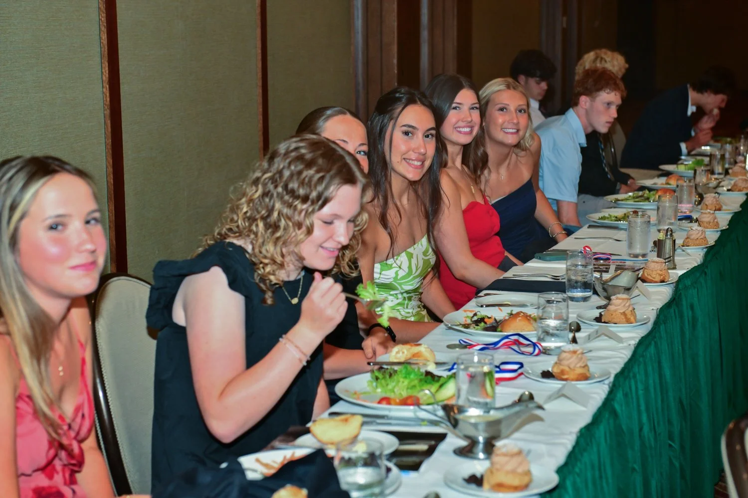 Group of young women and men sitting at a banquet table enjoying a meal at a formal event.