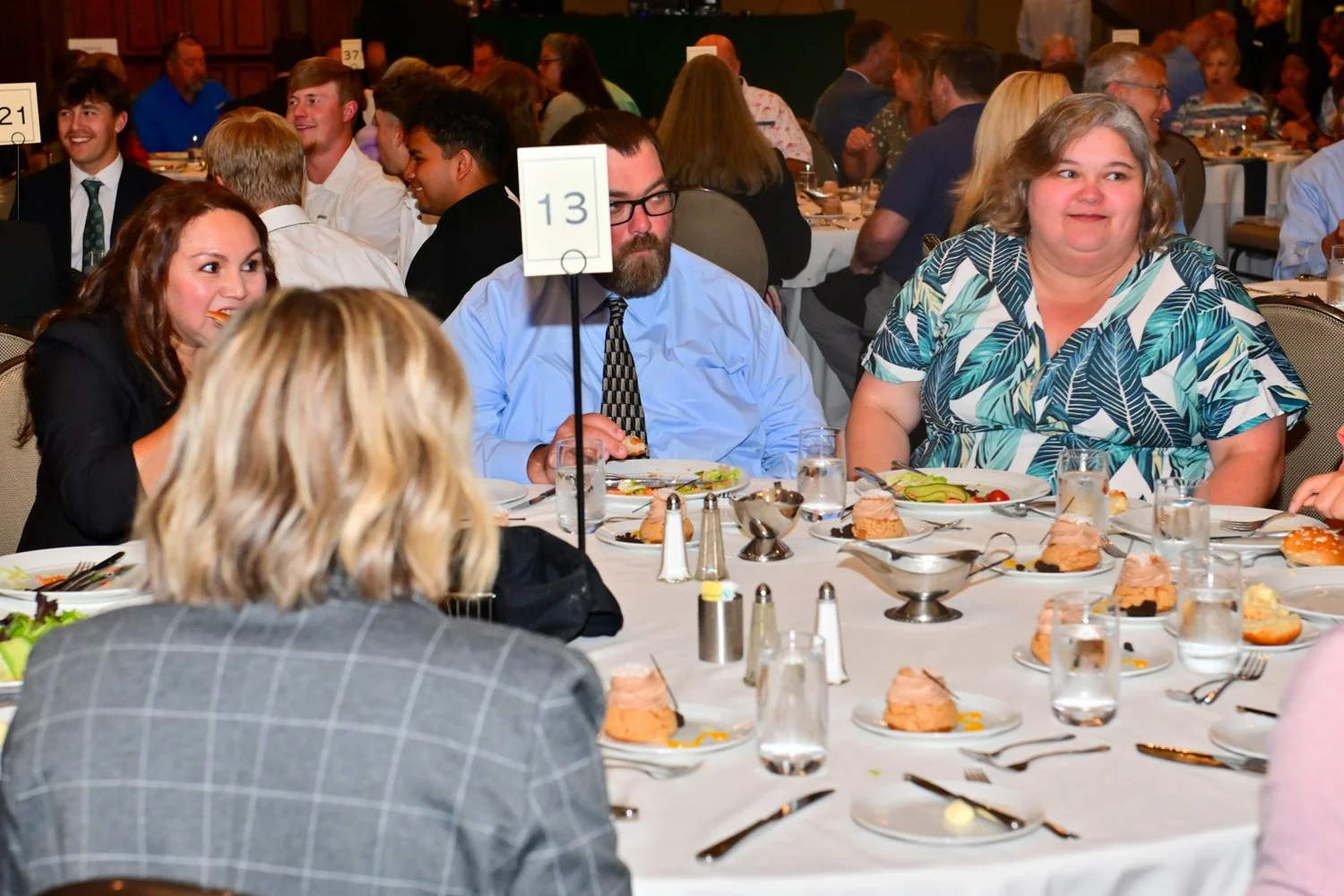 People sitting at a banquet table during a formal event, eating and engaging in conversation, with plates of food and drinks on the table.