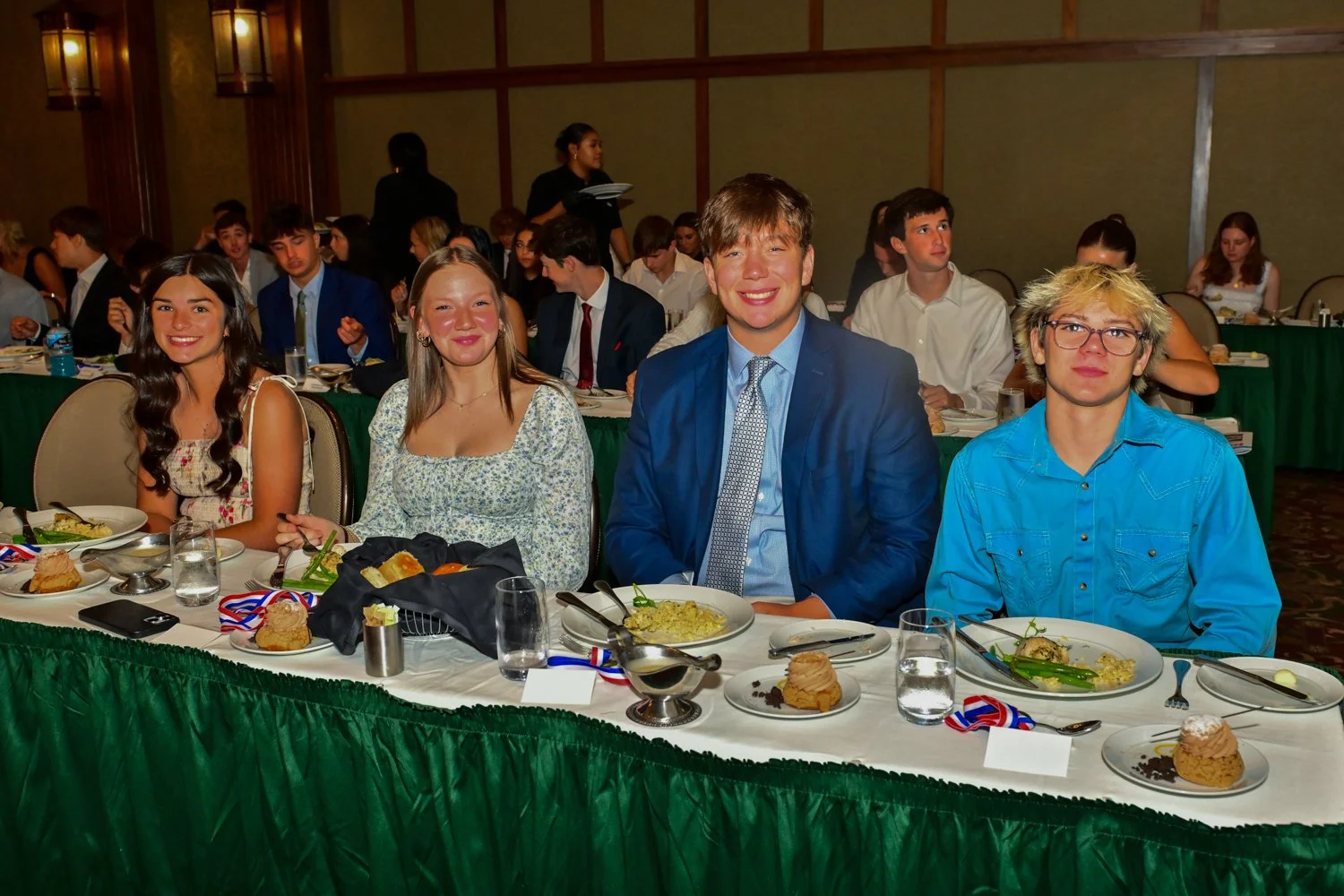 Group of four young people sitting at a banquet table with plates of food, drinks, and medals at an event, smiling at the camera.