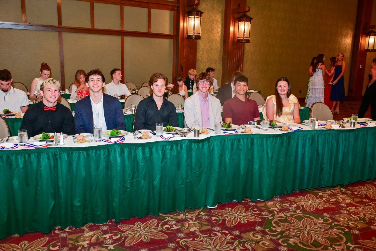 Group of seven young people seated at a banquet table with green tablecloth and plates of salad and bread, in a formal event hall with other guests in background.