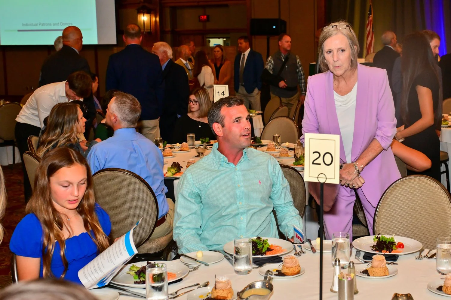 A woman in a lavender suit standing next to a seated man in a light green shirt at a banquet table during a formal event.