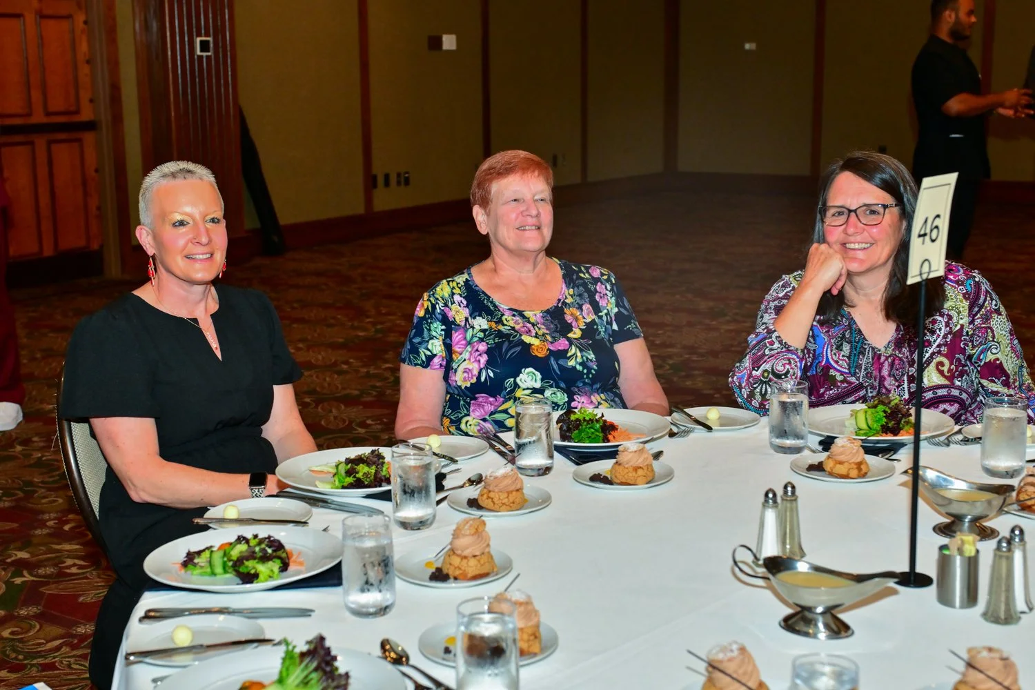 Three women seated at a round banquet table, smiling, with salads and desserts in front of them.