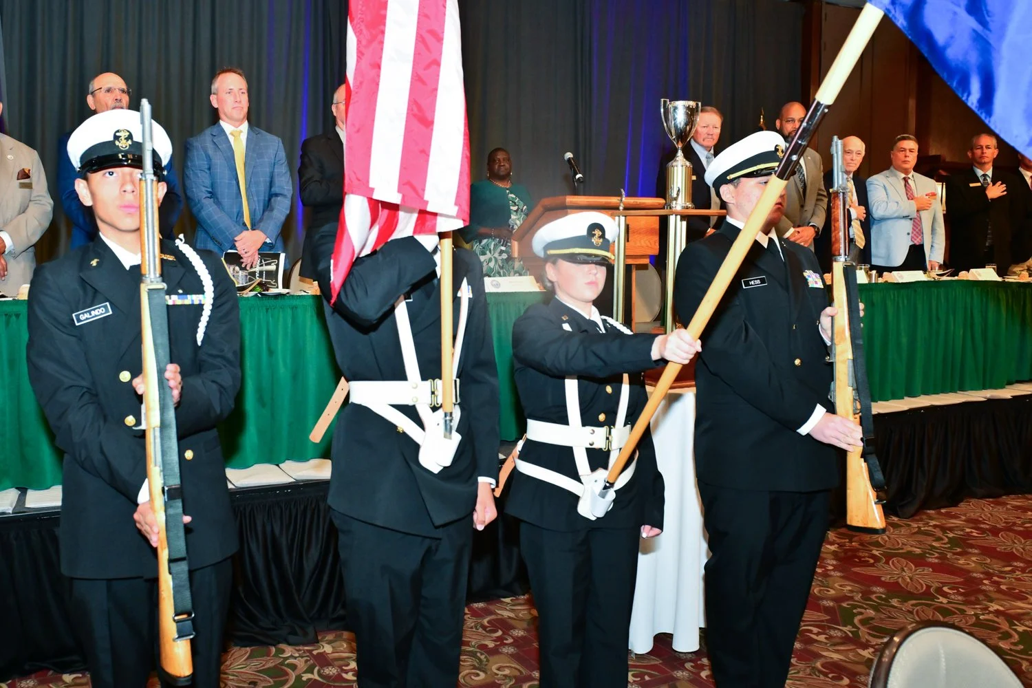 Four individuals in military uniforms holding rifles and flags during a formal ceremony, with a panel of officials seated at a long table behind them.
