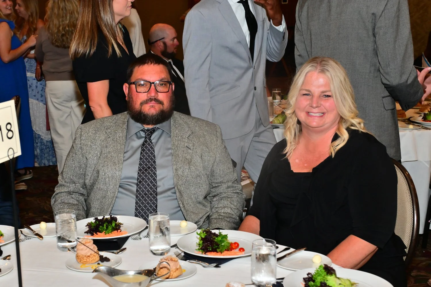 Two people sitting at a banquet table, smiling at the camera, with other guests in the background at an event.