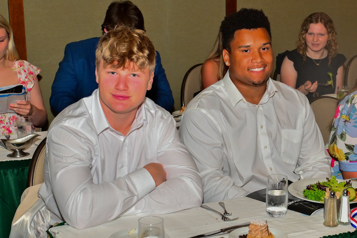 Two young men in white shirts sitting at a banquet table with food, drinks, and salad, smiling for the camera at a social event.