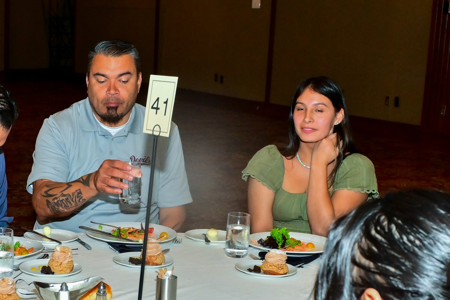 Two people sitting at a table with plates of food and drinks, woman in green top touching her neck, man in light blue shirt holding a glass.