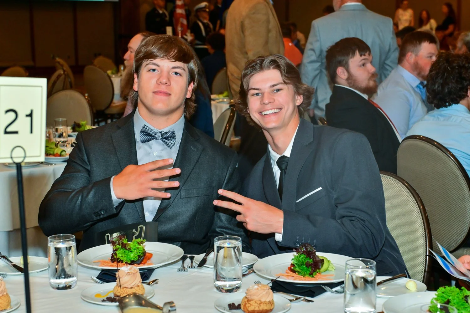 Two young men in suits sitting at a formal dinner table, smiling and making a peace sign. The table is set with plates of salad, drinks, and desserts.