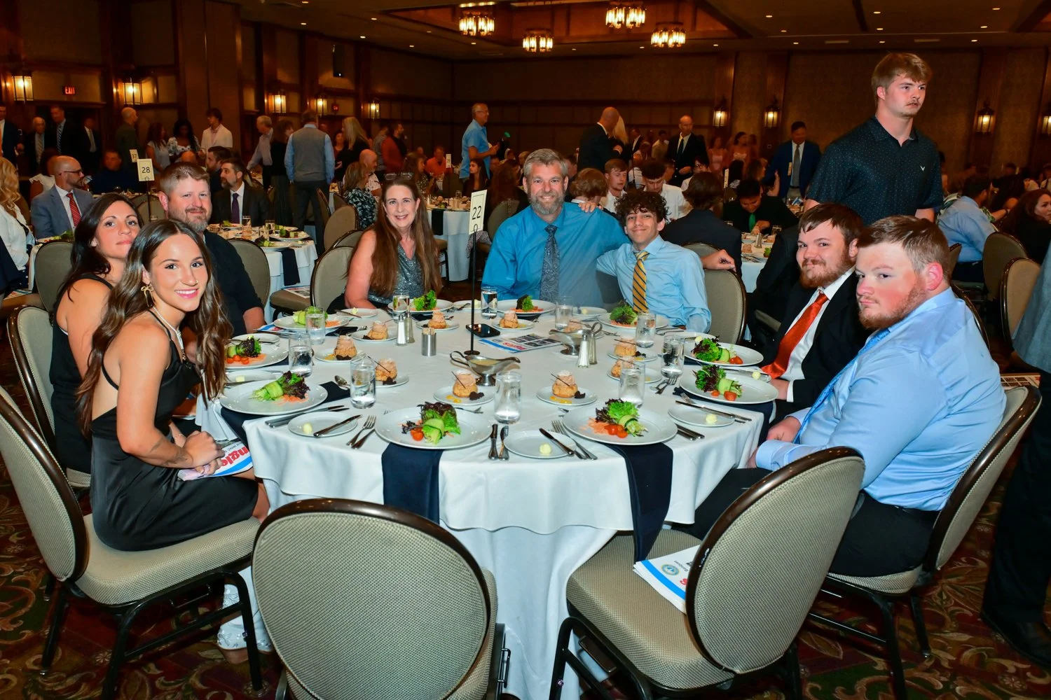 Group of people sitting around a round dining table at a formal event in an elegant banquet hall, with plates of food and glasses of water on the table.