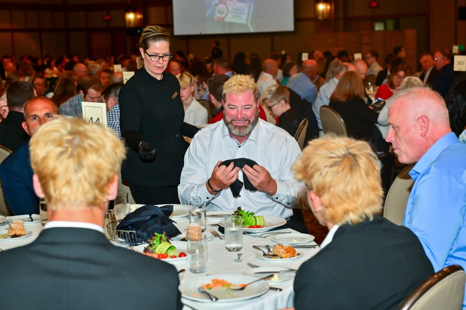 A man with gray curly hair and a beard smiling, holding glasses, seated at a round banquet table during a large formal event, with other guests and a waiter in black uniform in the background.
