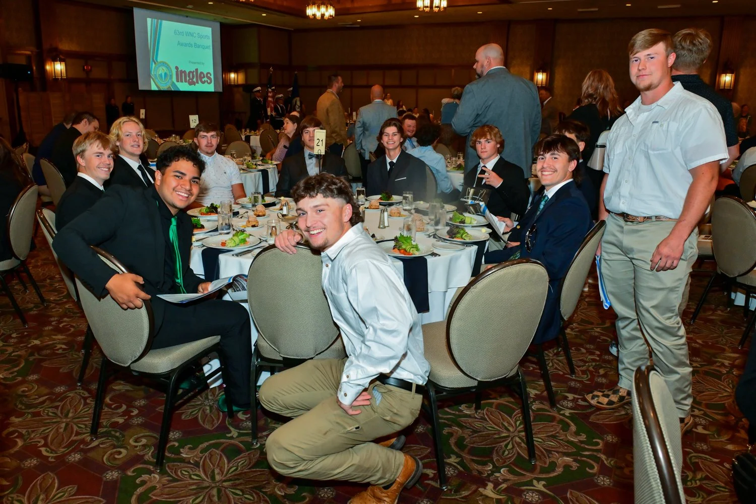 Group of young people dressed in formal attire, seated at a round banquet table, attending a formal event or award banquet in a decorated banquet hall.