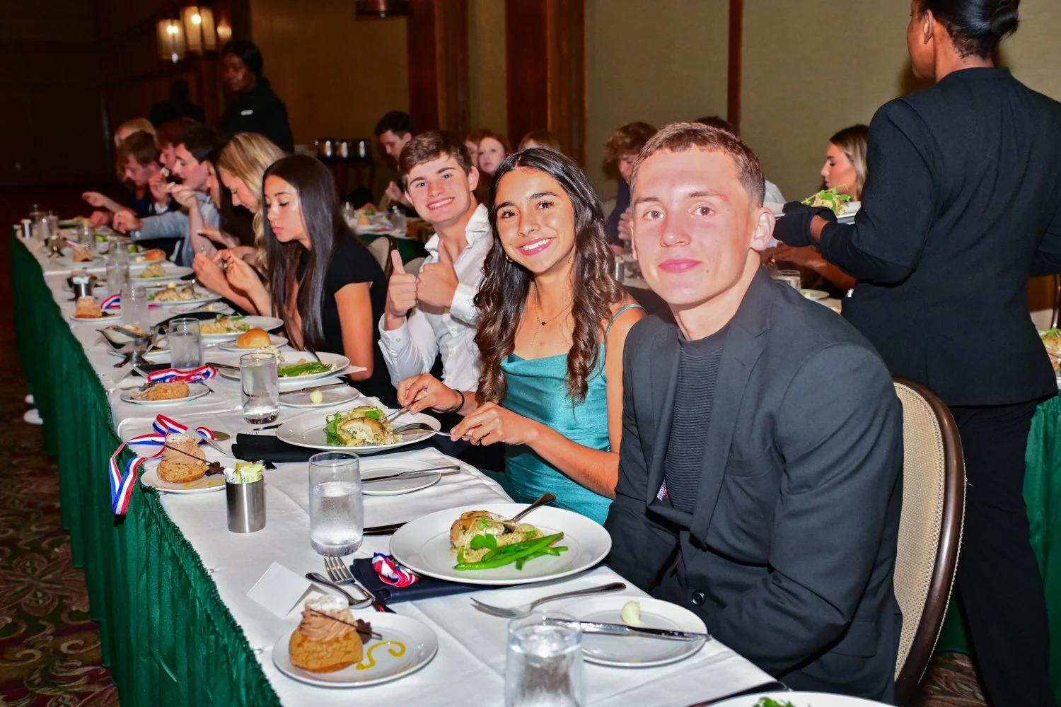 Young people sitting at a banquet table during a formal event, eating and smiling, with a few giving thumbs-up signs, dressed in formal attire with a green tablecloth and decorated plates.