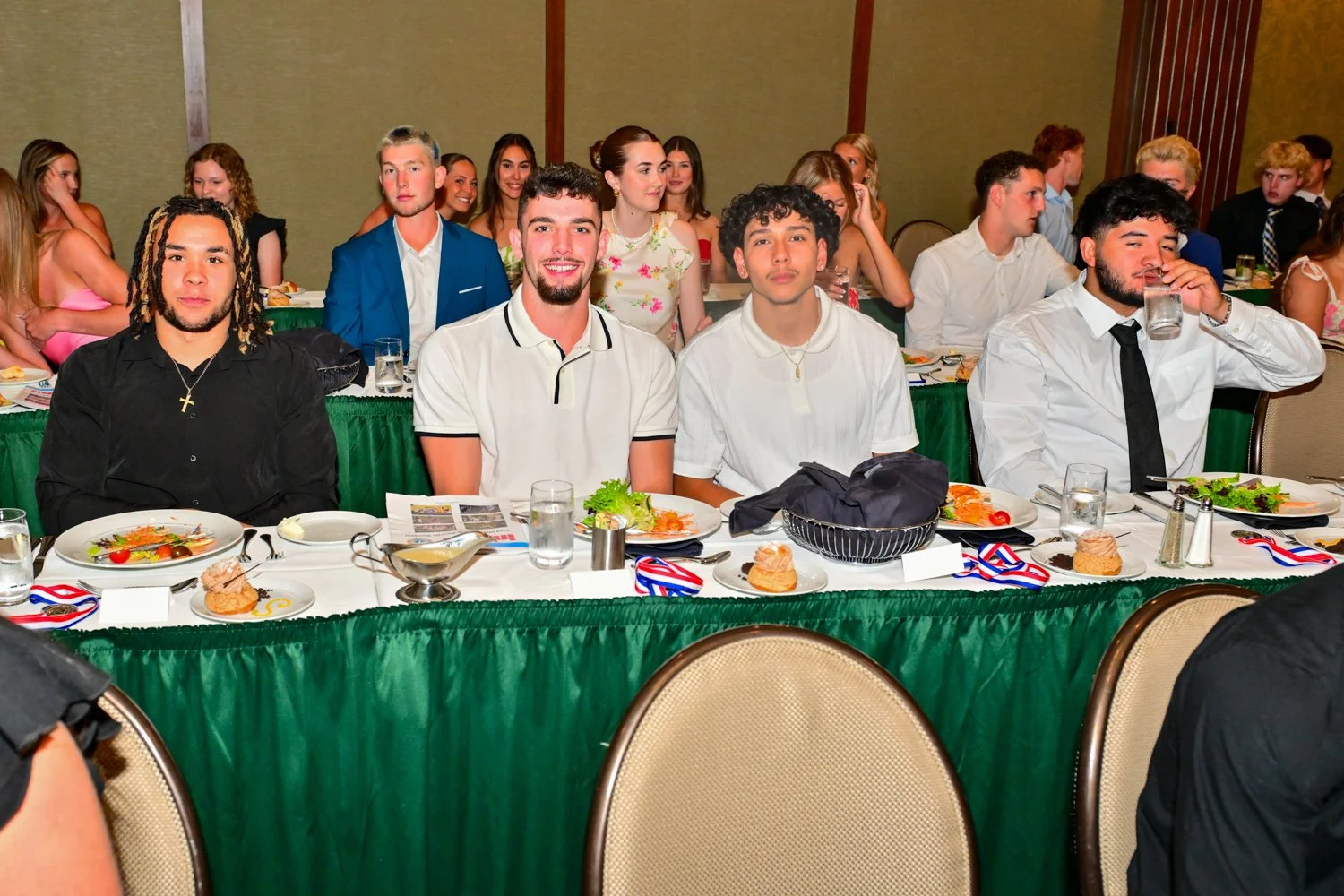 Group of young people sitting at a banquet table during a formal event or celebration, with some eating and others engaging in conversation.