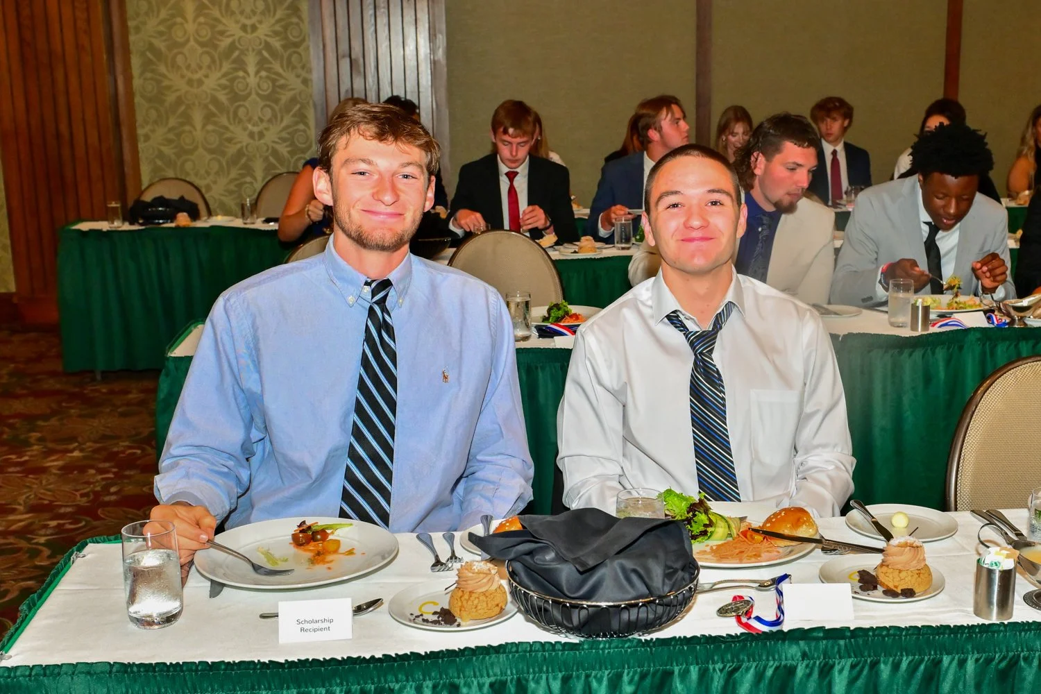 Two young men in dress shirts and ties sitting at a banquet table with food and desserts, smiling at the camera, during a formal event.
