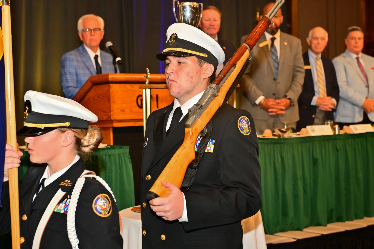 A young man in a black military uniform and white hat is holding a rifle during a formal ceremony. A young woman in a similar uniform is partially visible beside him. In the background, several men in suits are standing behind a green-draped table, w