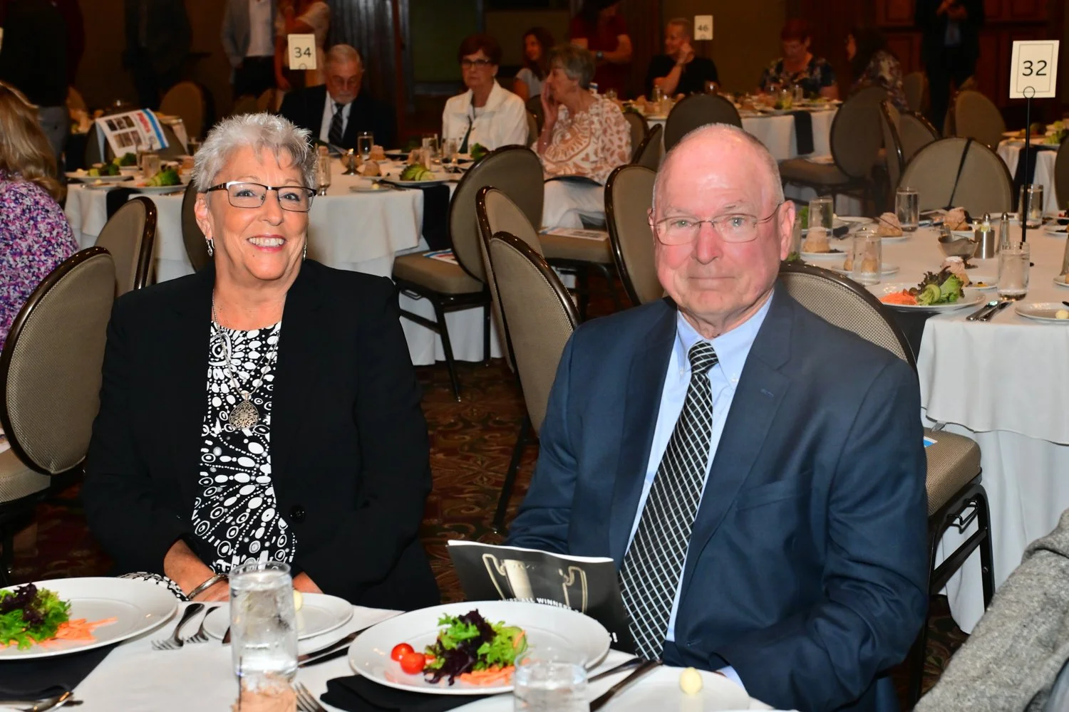 A woman and a man sitting at a table during a formal event or dinner, with other guests seated at tables in the background.