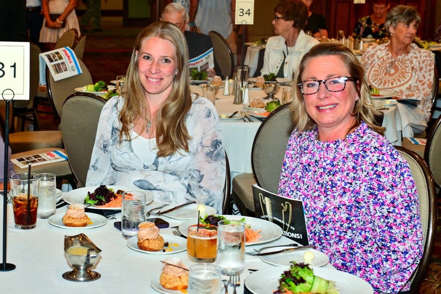 Two women sitting at a table during a formal event. The table is decorated with plates of food, drinks, and desserts. In the background, other guests are seated at tables and dressed in semi-formal attire.