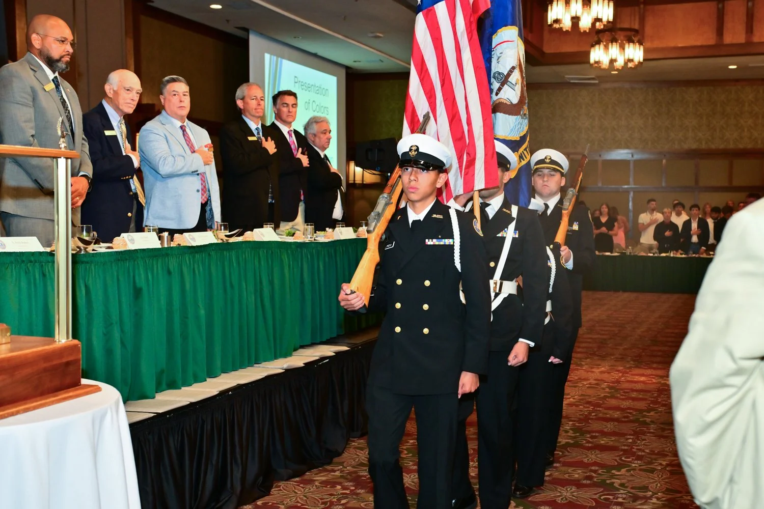 A color guard procession with young military personnel holding the U.S. flag and military flag, marching in a formal event at a banquet hall with officials and attendees watching.