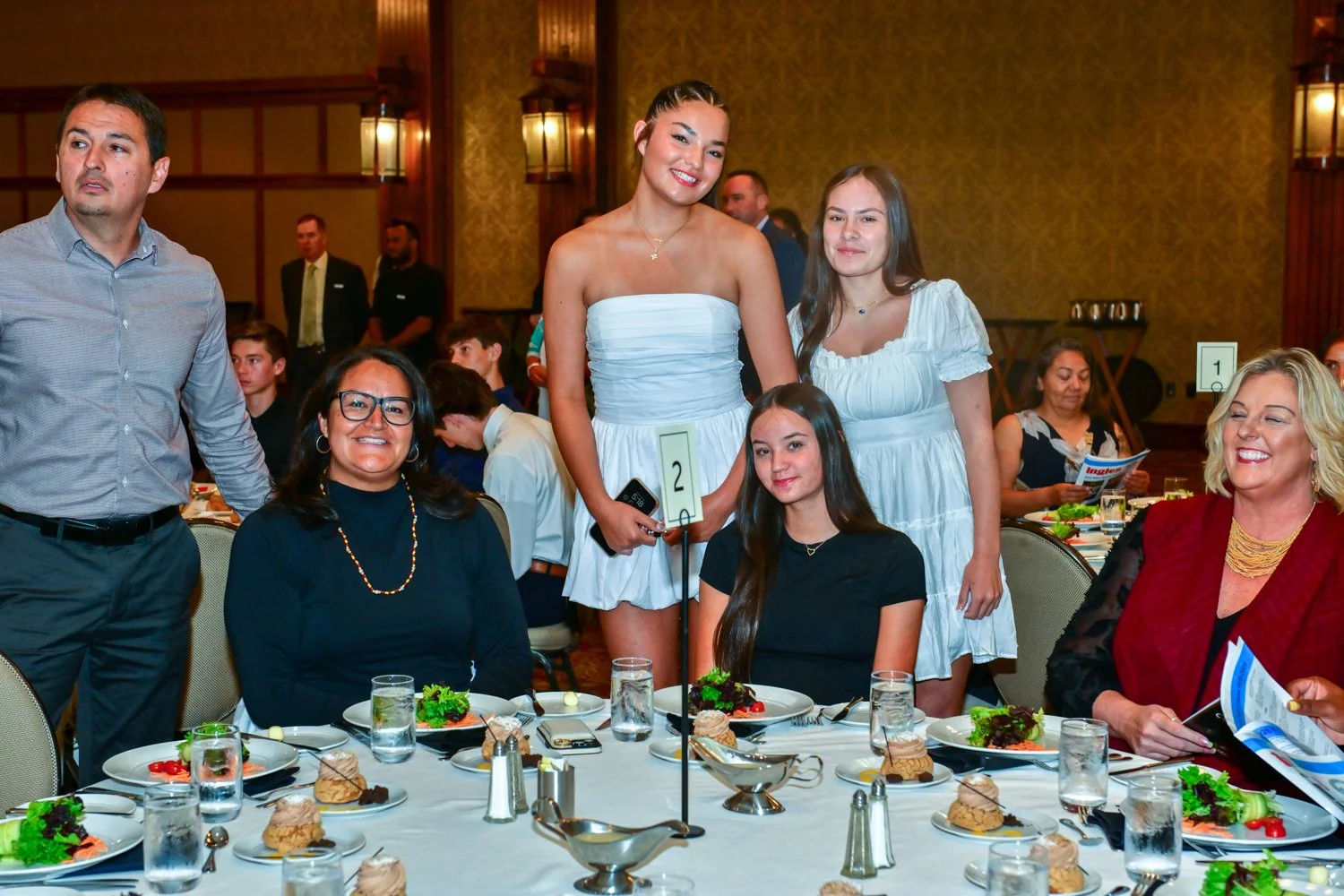 Group of six people, five women and one man, gathered around a dining table at a formal event. Four women are seated, and two women and one man are standing behind them. The table is set with plates, salads, desserts, and drinks.