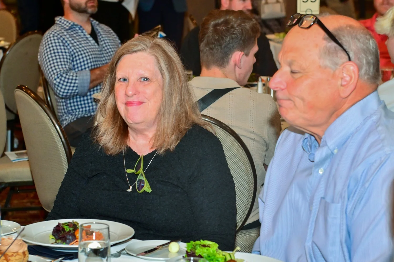 A woman with shoulder-length light brown hair and a black top, smiling, sitting at a table with a woman and man in a lively gathering or banquet setting.