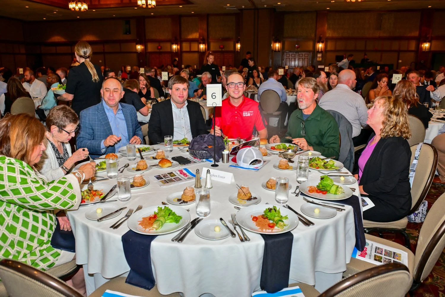People sitting around a banquet table at a formal event, with plates of salad, bread, drinks, and a centerpiece with a table number. The room is filled with other guests at similar tables.