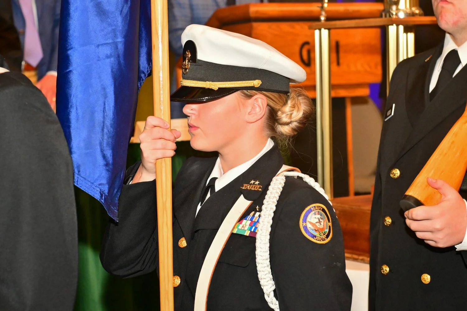 A female U.S. Navy officer in uniform holding a flag at a formal ceremony.