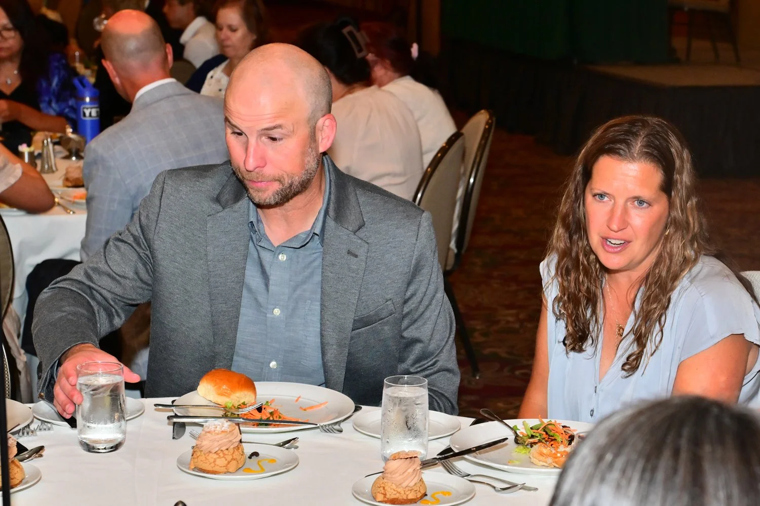 People sitting at a banquet table with food, including a man in a gray suit and a woman with curly light brown hair, at a formal event or dinner gathering.