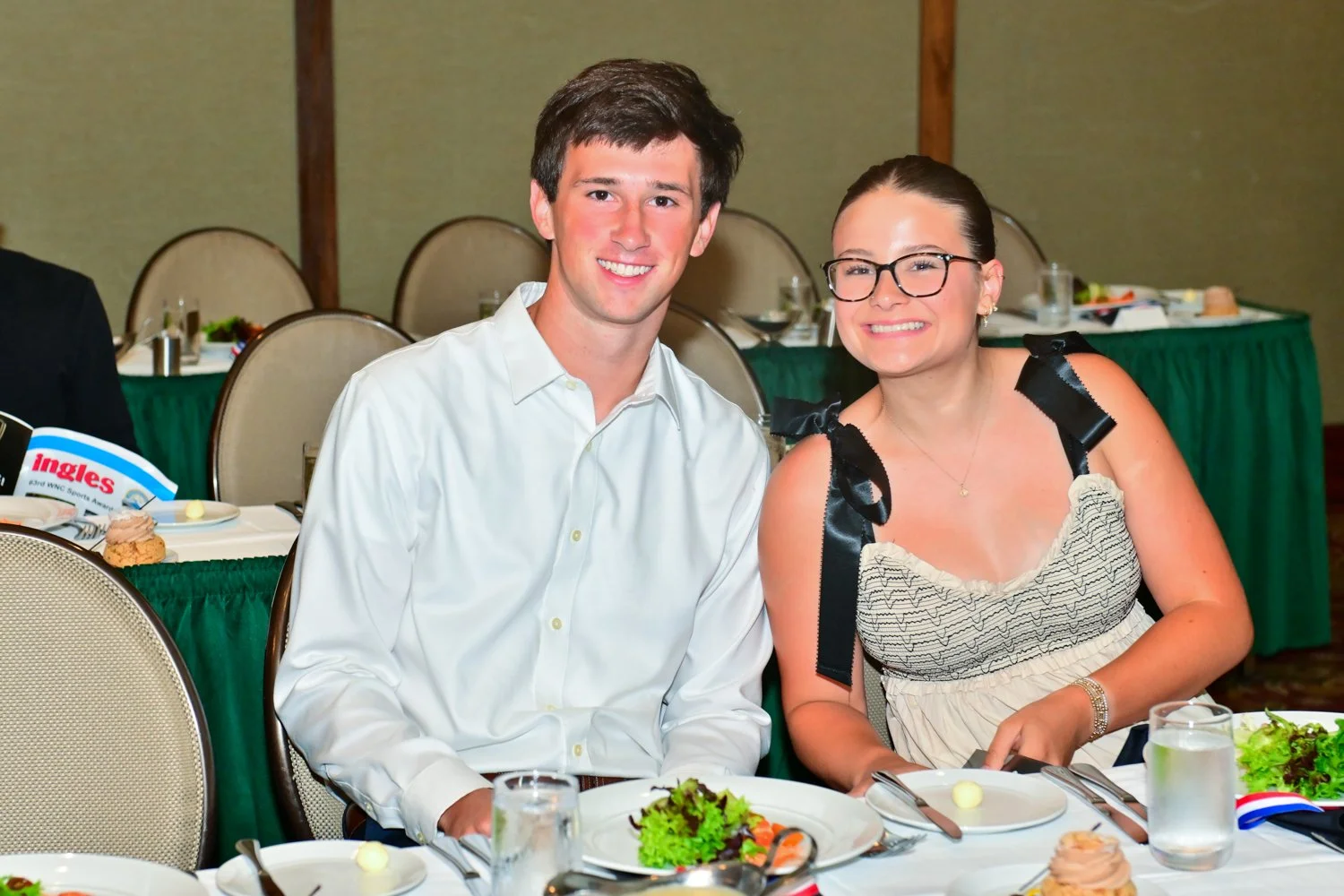 A young man and woman sitting at a table during a formal event. They are smiling at the camera, with the man in a white shirt and the woman wearing glasses and a sleeveless dress. The table is set with plates, utensils, and drinks, with other tables 