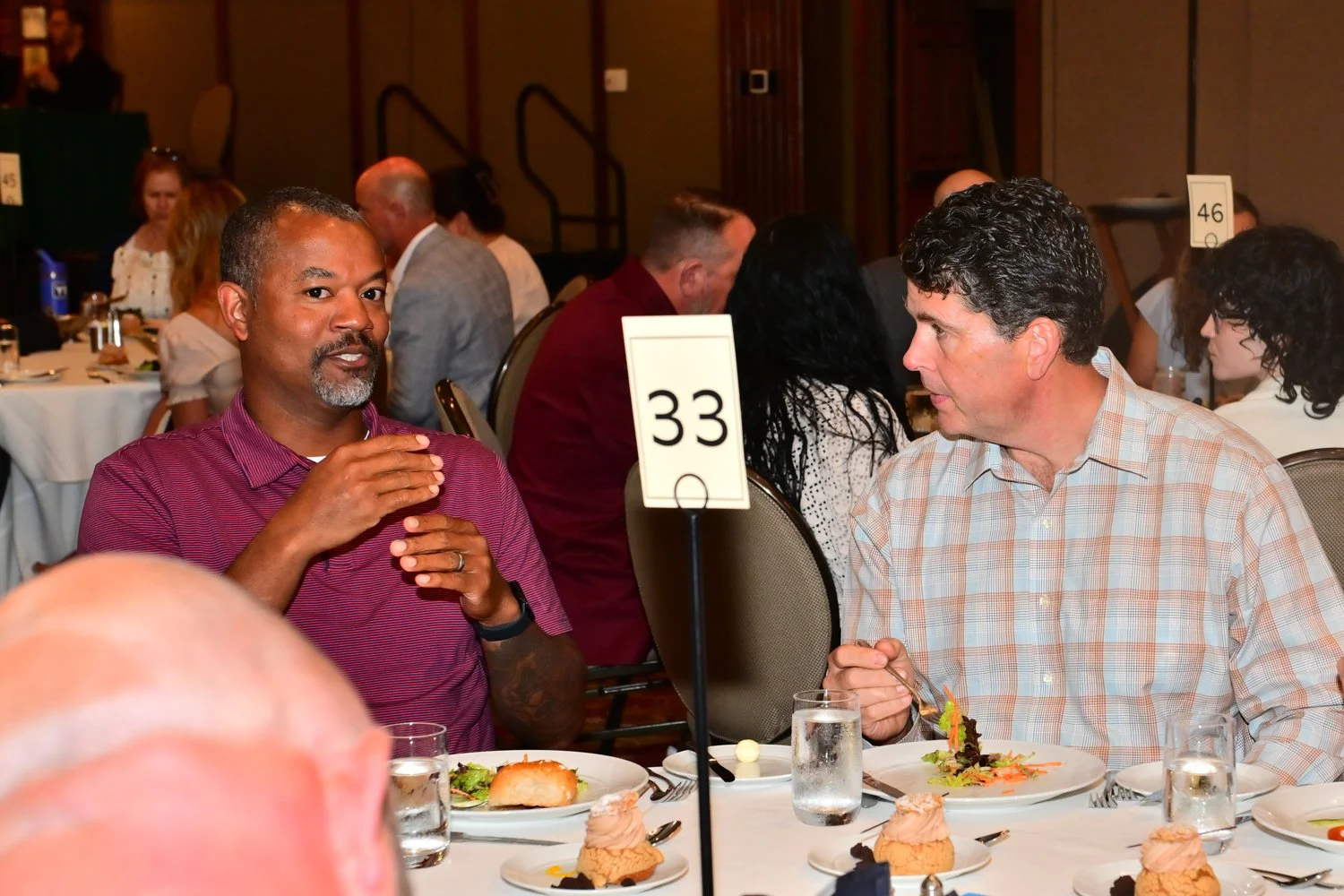 Two men sitting at a banquet table engaged in conversation, with plates of food and desserts in front of them, at a formal event.