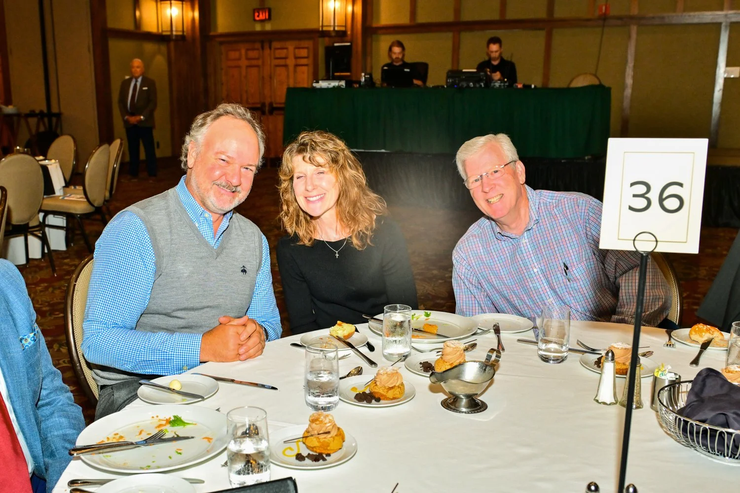 Three people sitting at a round banquet table with dessert, glasses of water, and cutlery, smiling. A man in a gray vest, woman with curly hair, and man with glasses wearing a checkered shirt. In the background, two DJs and a man standing with hands 