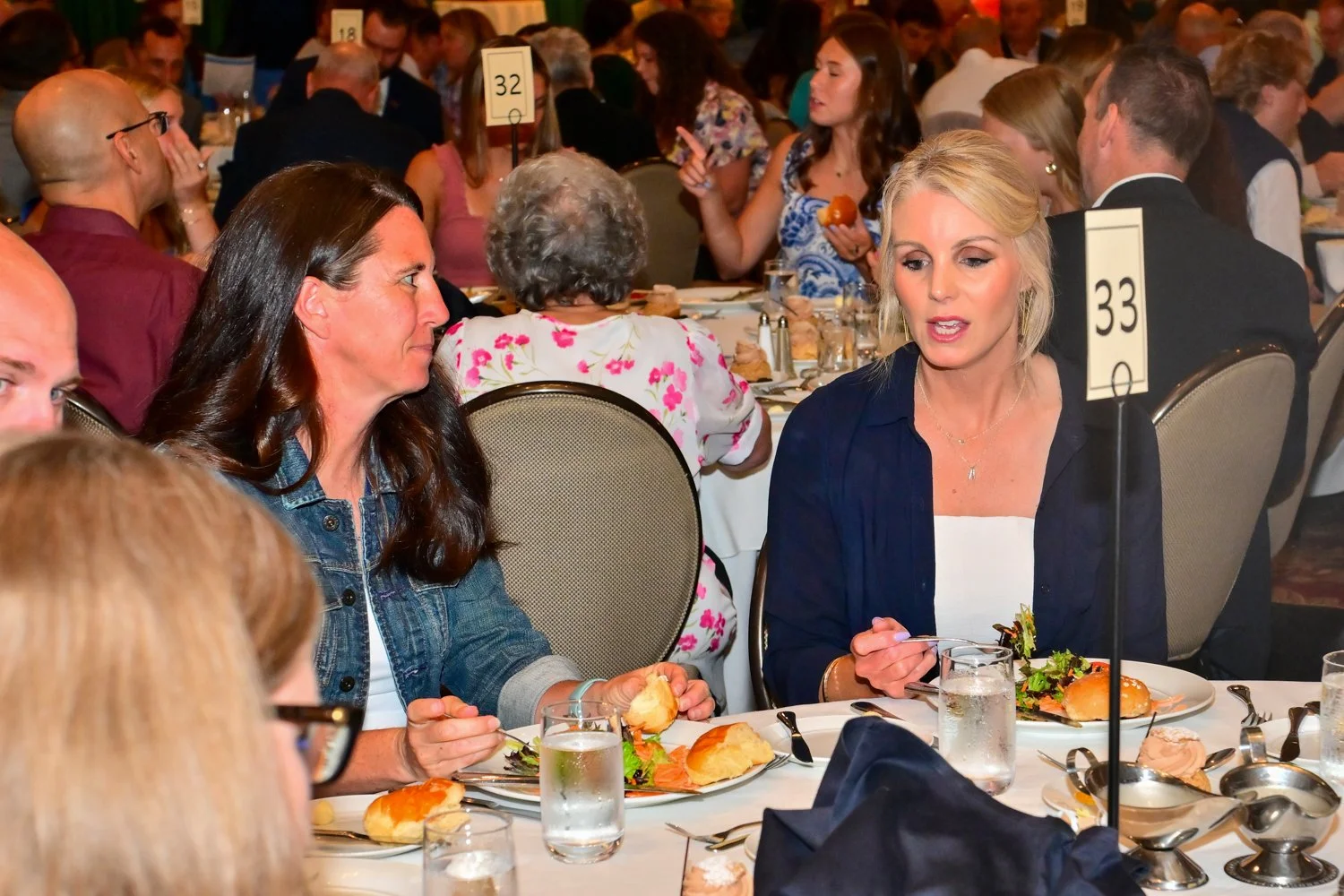 People seated at a banquet table during an event, eating and talking, with table number 33 visible, and plates of food including sandwiches and salads.