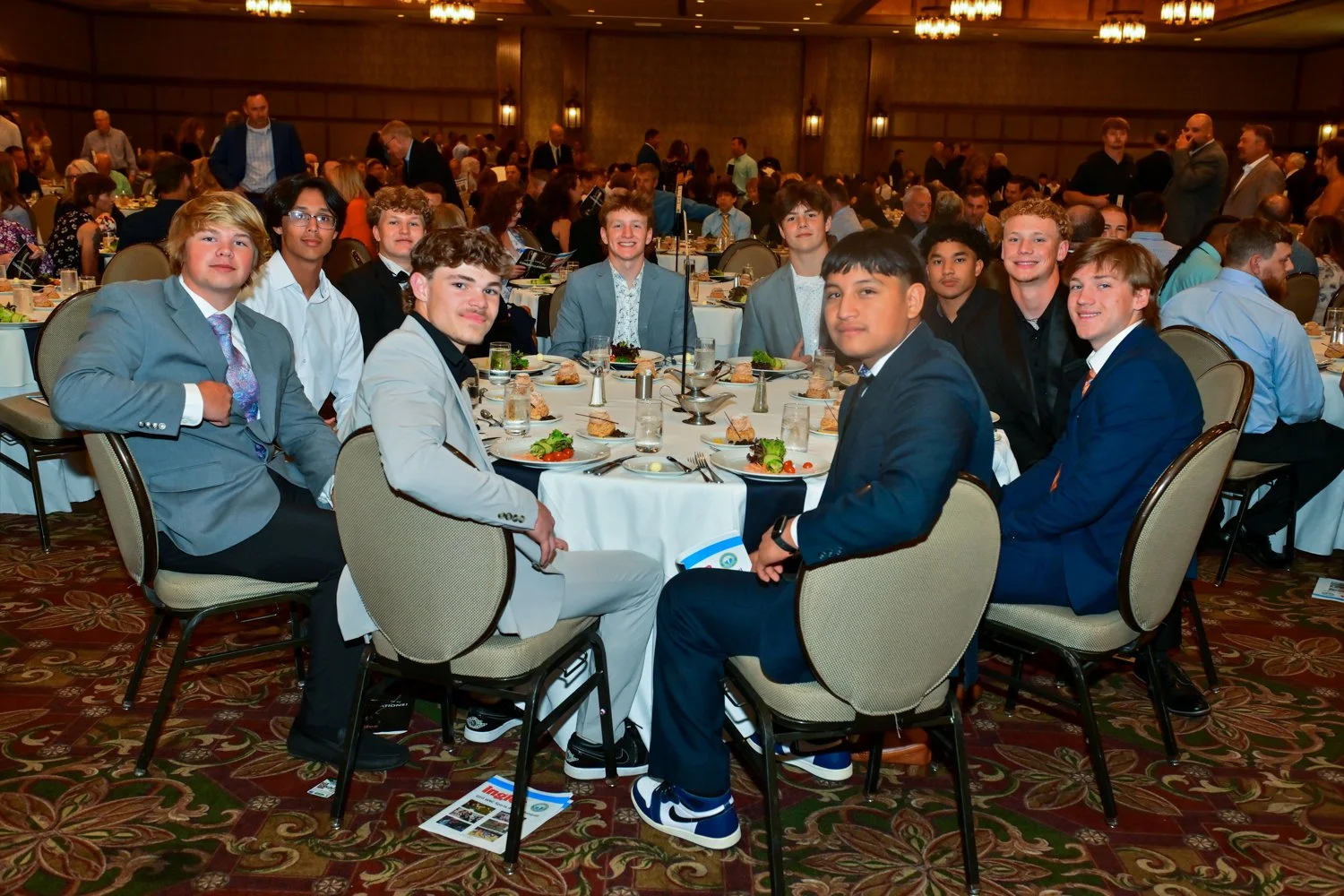 A group of young men in formal suits seated around a dining table at a banquet hall with many other guests in the background.