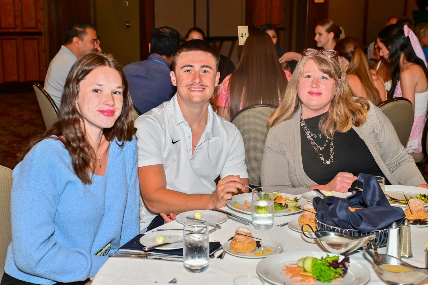 Three people sitting at a dinner table during a formal event, with other guests in the background. The table has plates of food, glasses of water, and utensils.