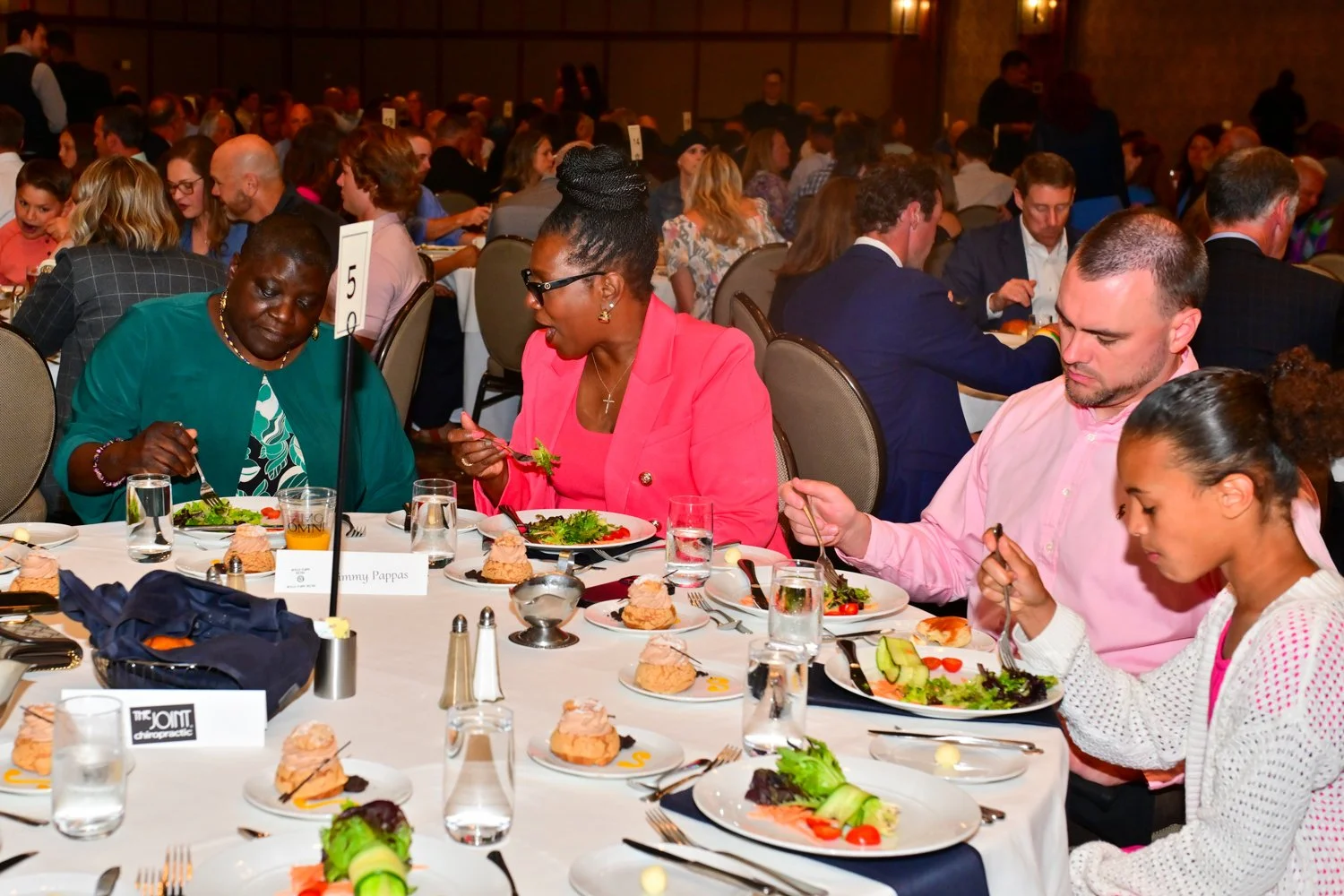 People dining at a formal event with multiple tables, plates of food, and glasses of water and juice.