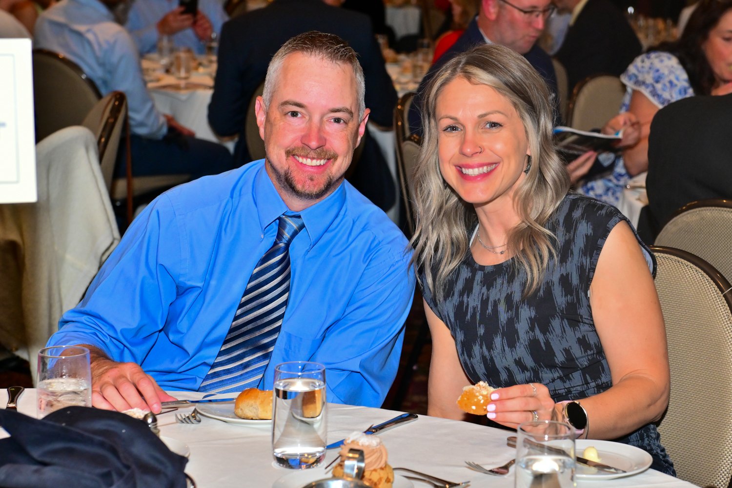 A smiling man and woman sitting at a banquet table during a formal event, with glasses of water, a bread roll, and a dessert on the table.