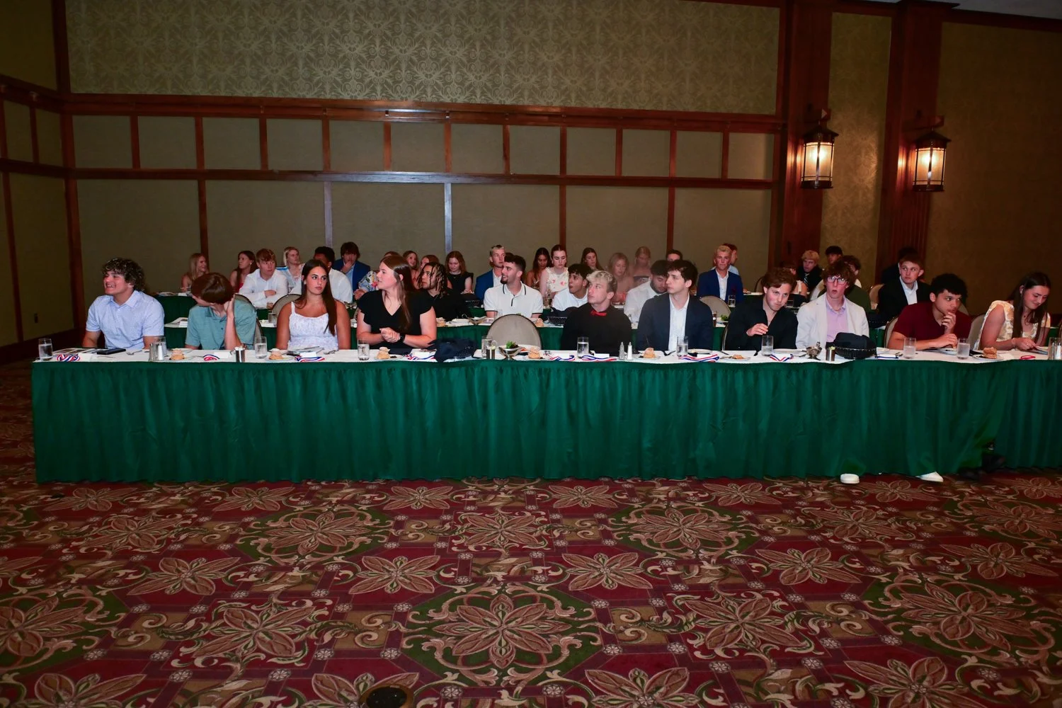 A group of young adults seated at long tables during an event or conference in a large, ornate room with green and gold wallpaper and wooden accents, with a patterned carpet on the floor.