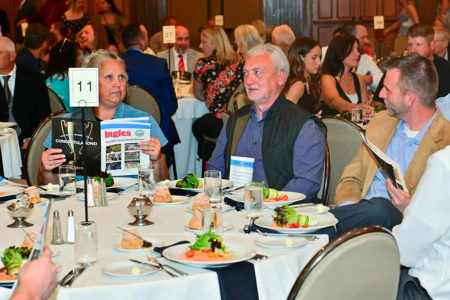 People sitting around a banquet table with plates of salad, bread, and glasses of water, attending an awards banquet, with woman holding a magazine and conversation among guests.