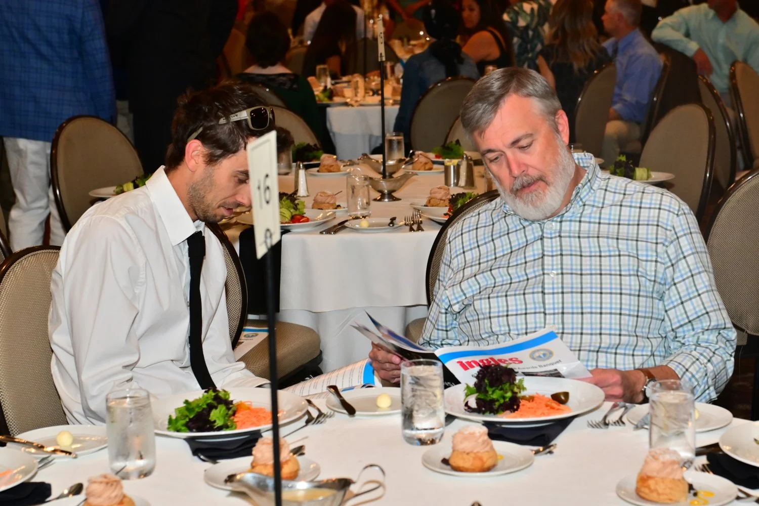 Two men sitting at a banquet table, looking at a magazine. The table is set with plates of salad, glasses of water, and small desserts. The background shows other people seated and standing at similar tables.