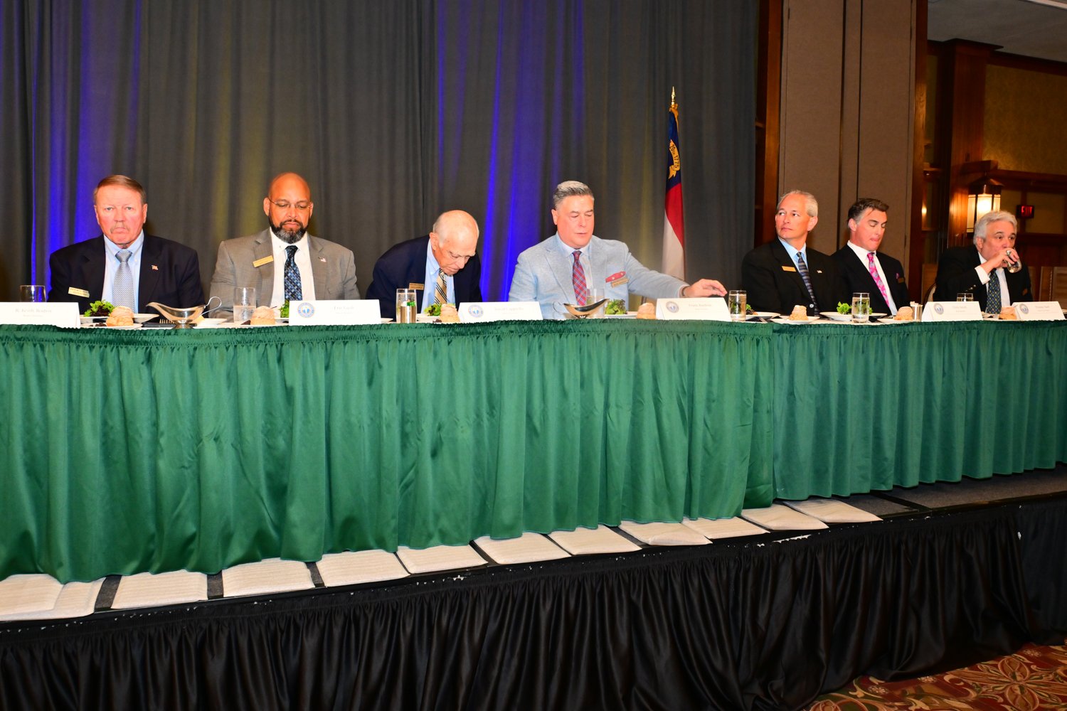 A group of eight men sitting at a long table on a stage during a formal event, with a blue curtain backdrop, a flag, and water glasses on the table.