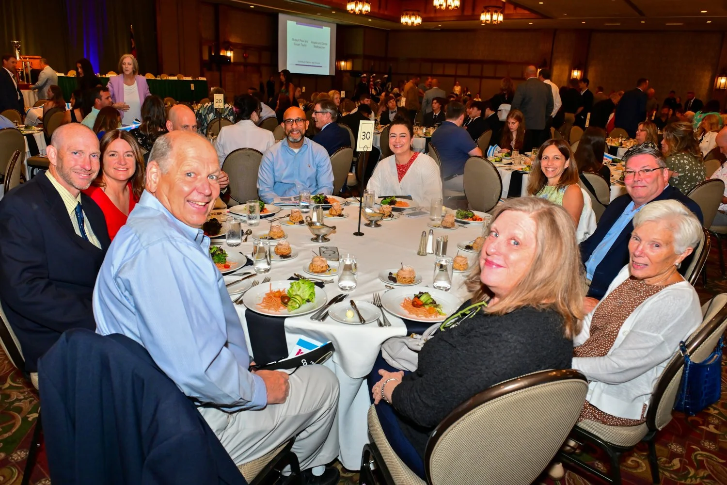A group of people gathered around a round banquet table at a formal event, with plates of food, drinks, and silverware, smiling at the camera in a decorated banquet hall.