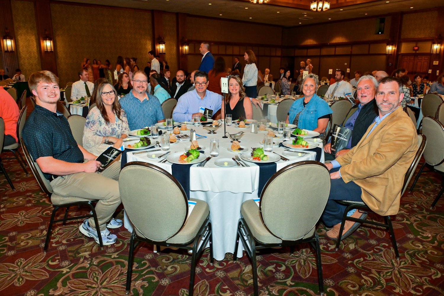 A group of nine people sitting around a banquet table at a formal event in a large decorated hall, with other tables and guests in the background.