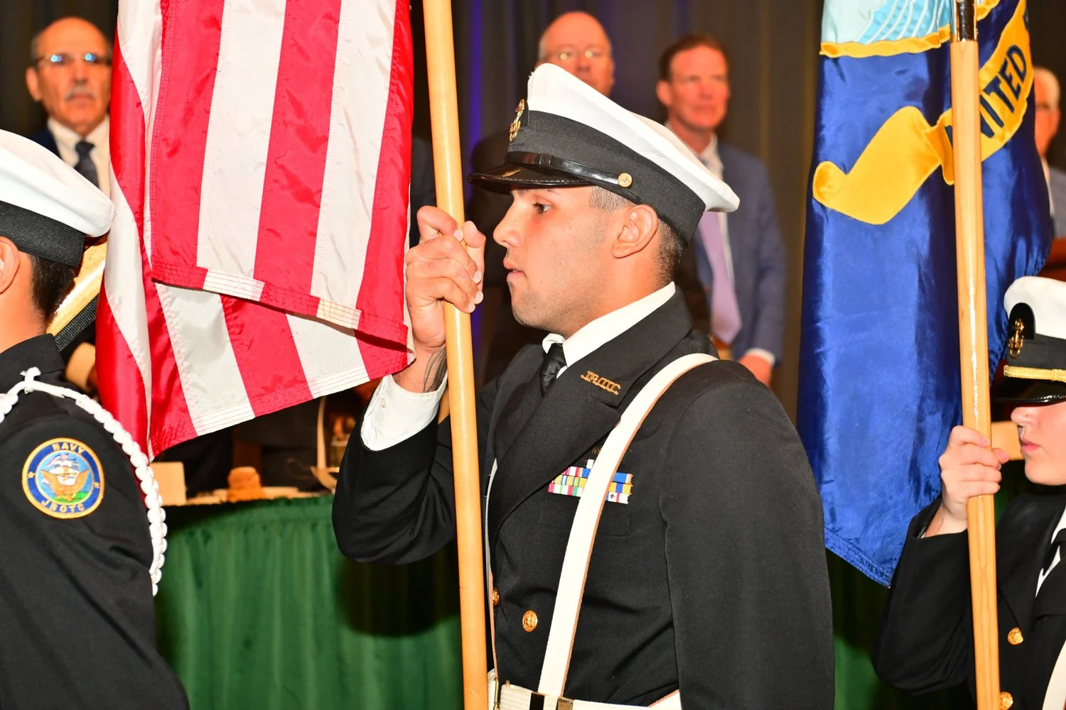 A uniformed man participating in a flag ceremony, holding a United States flag, with other flag bearers and attendees in the background.