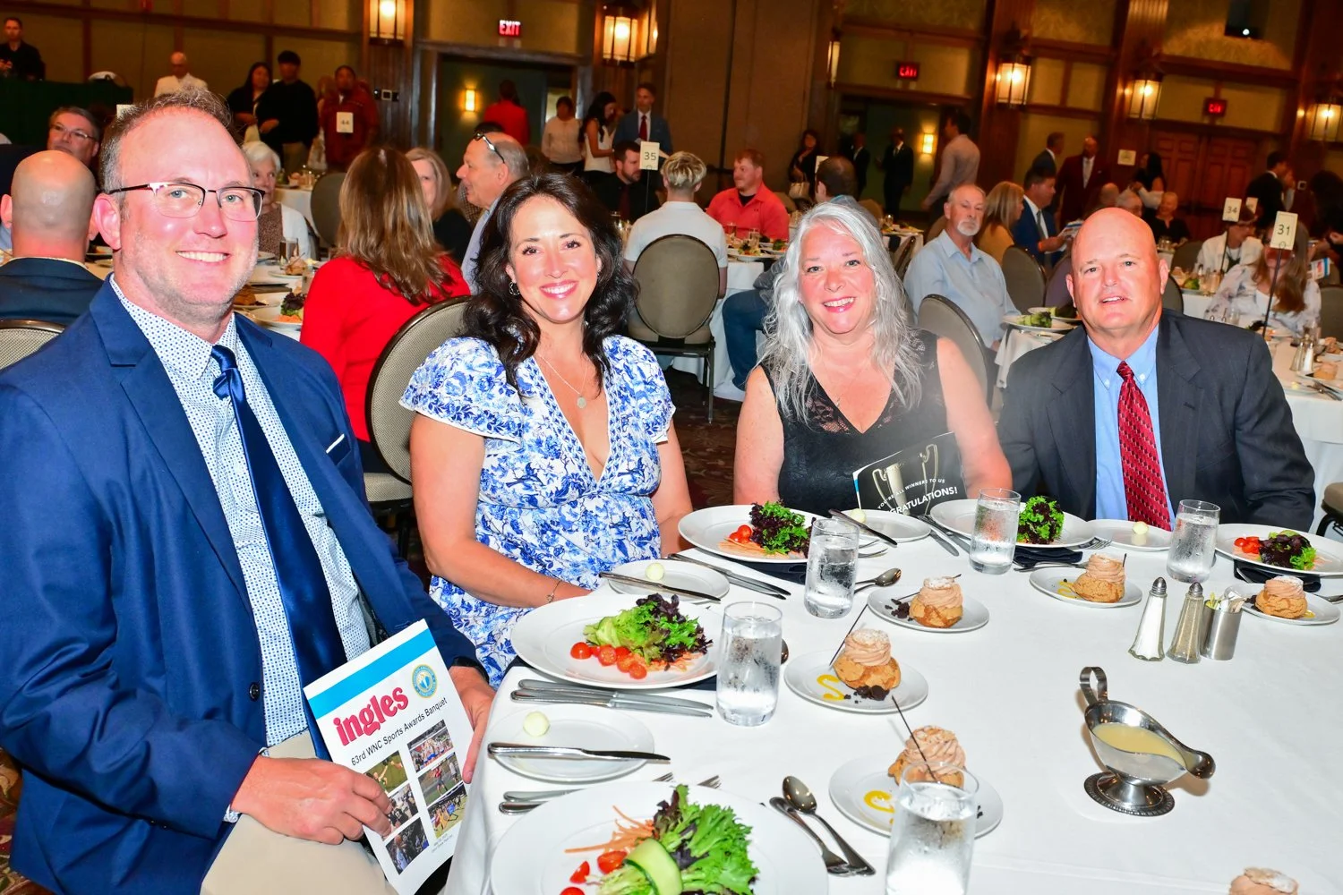 Group of four smiling people sitting at decorated banquet table with salads, desserts, and drinks, in a large event hall with other guests dining in the background.