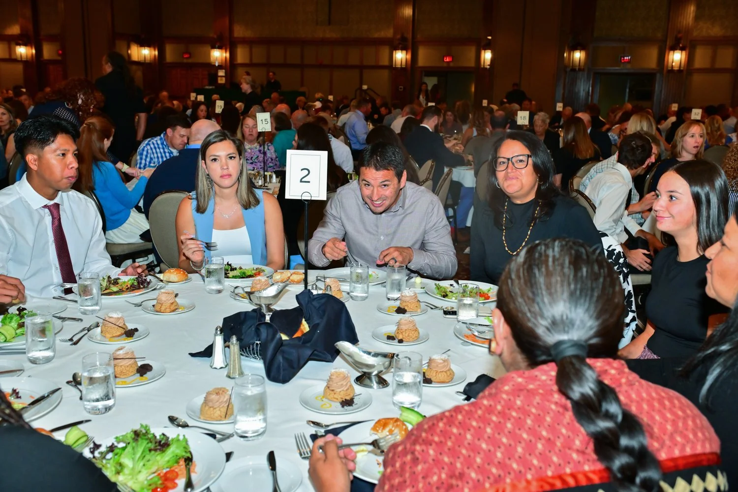People sitting at a round banquet table during a formal event, with plates of food and desserts, in a large decorated conference room.