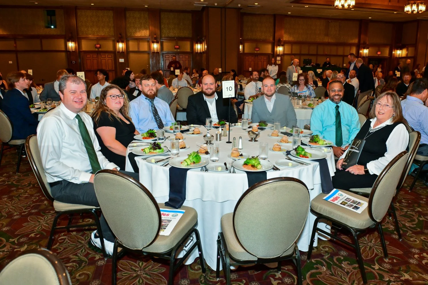 Group of people sitting at a round banquet table with white tablecloths, plates of salad and sandwiches, and a table number sign at a formal event or conference.