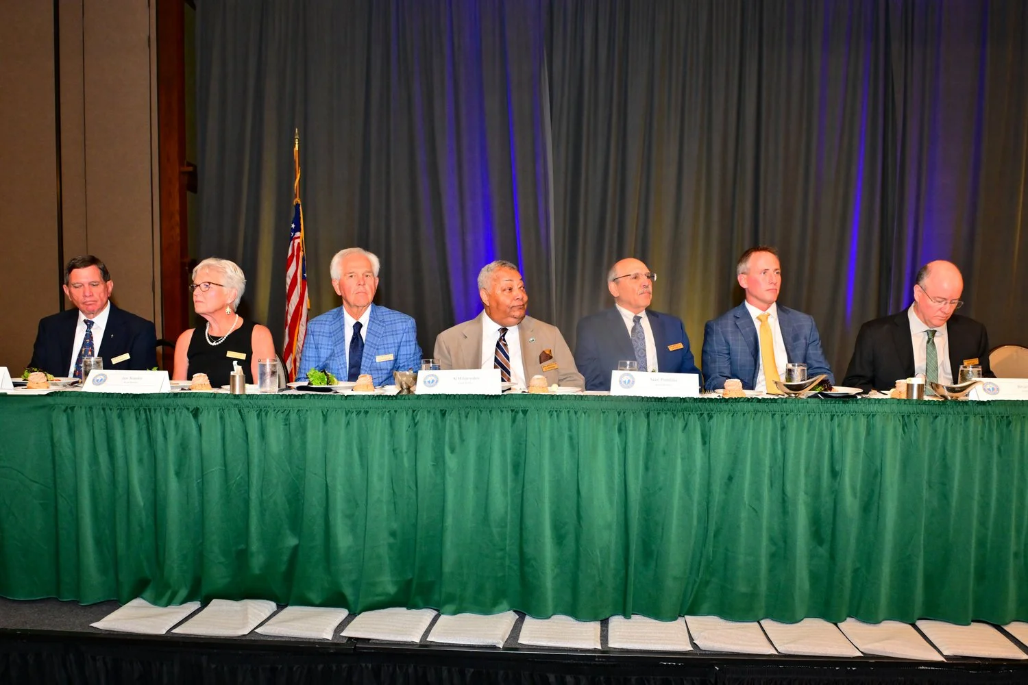 A group of eight people seated at a long green table with nameplates and microphones, in front of a dark curtain and an American flag, during a formal event or meeting.