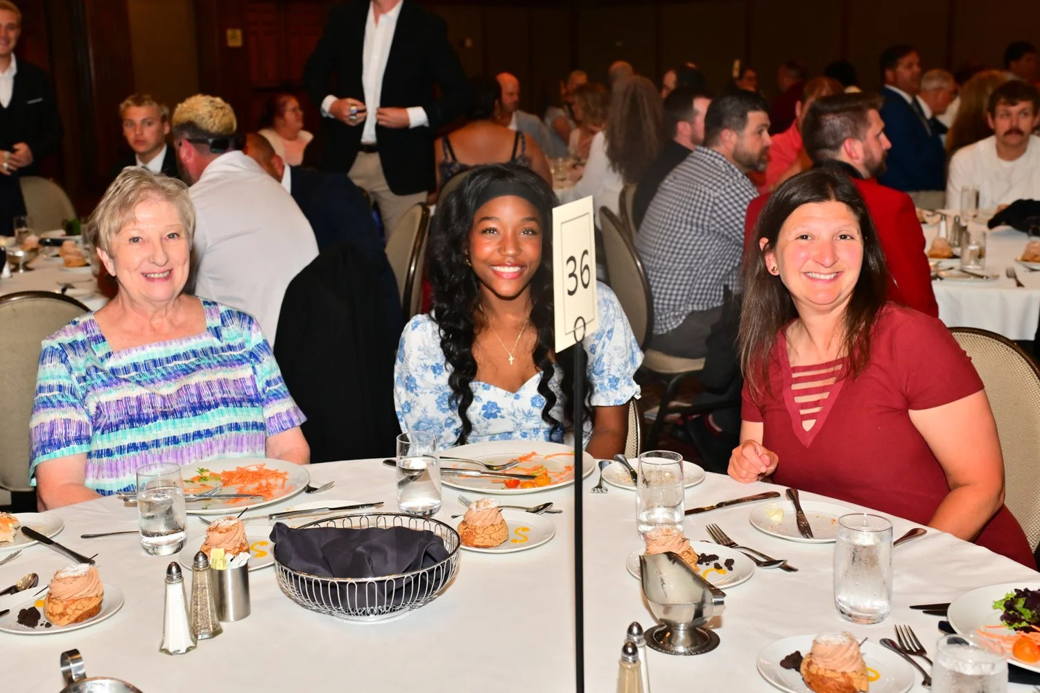 Three women sitting at a banquet table during a formal event, smiling for the camera. The table is set with plates, glasses, and desserts.
